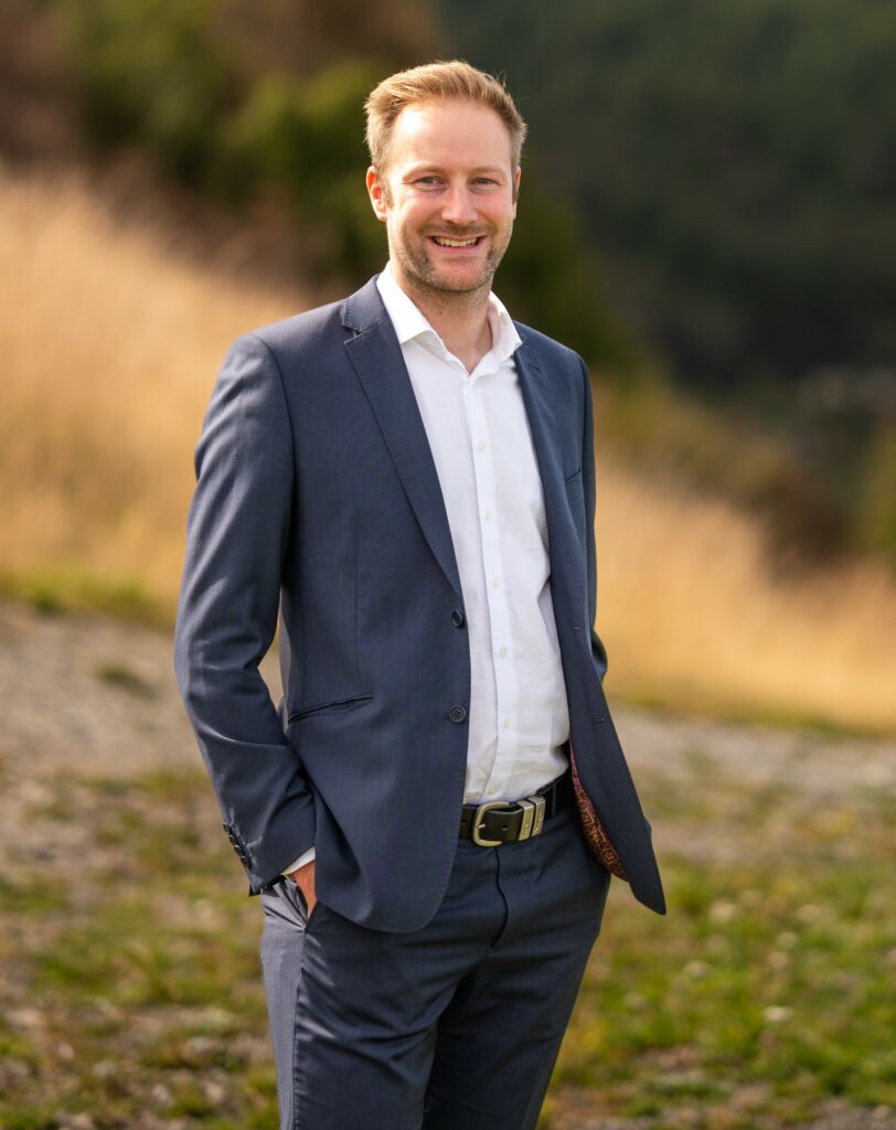 Jono in professional corporate attire with his hands in his pockets, standing outside with landscape behind