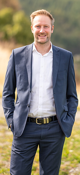 Jono in professional corporate attire with his hands in his pockets, standing outside with landscape behind